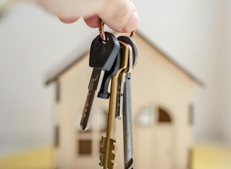 Close-up of a hand holding keys with a miniature wooden house in the background, symbolizing real estate investment.